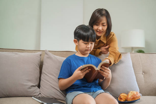 istock Asian mother and son sit together on a comfortable sofa at home, reading a book while watching a tablet, enjoying a peaceful and educational family time 2191019143