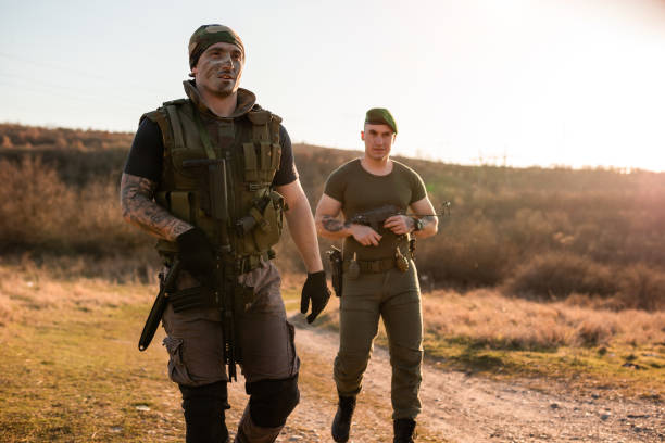 Two individuals in military gear performing tactical exercises in an open field during daylight. The scene depicts action and teamwork, emphasizing outdoor training activities in a strategic environment suitable for military or survival scenarios.