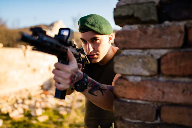 A focused soldier wearing a green beret takes aim with a rifle while positioned behind a brick wall, illustrating tactical defense and precision outdoors during daylight.