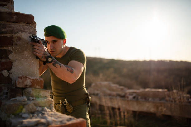 A focused soldier in green beret aims rifle through a broken wall, outdoors during daylight.
