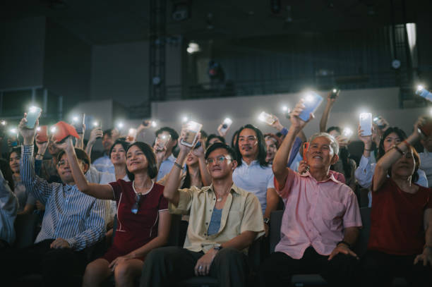 Asian senior man waving phone with flashlight singing along with Crowd  At Concert