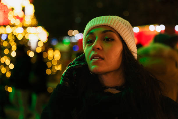 A young woman, illuminated by the vibrant holiday lights, speaks on her phone while enjoying the atmosphere of the Berlin Christmas market. Her white beanie and black gloves highlight her winter style amidst the golden bokeh of Christmas decorations in the background