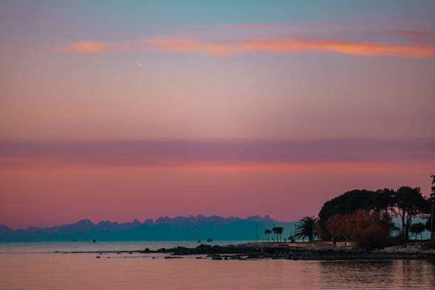 A vibrant sunset over a calm coastal scene, featuring pastel hues of pink, purple, and blue in the sky. Silhouettes of trees and a peaceful shoreline frame the tranquil water, with distant mountains adding depth to this serene landscape.