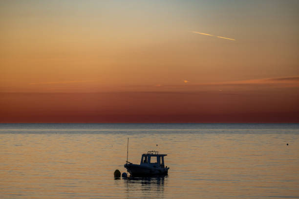 A peaceful scene of a small boat floating on calm ocean waters during sunset. The sky transitions through warm hues of orange and red, creating a serene and tranquil atmosphere. This picturesque setting captures the beauty of nature and solitude.