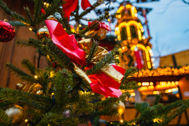 A close-up of a beautifully tied red and gold ribbon on a Christmas tree branch at the Berlin Christmas market. The warm bokeh lights in the background create a magical holiday ambiance, reflecting the joy and festivity of the season