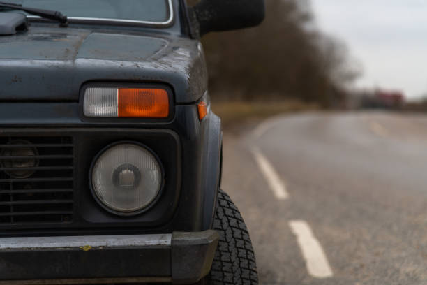 Close-up front view or round headlight of old gray coloured dirty and rusty car standing outdoors on autumn or spring countryside road. Soft focus. Copy space. Automobile details theme.