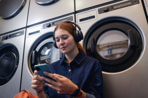 istock Girl Using Phone and Listening to Music while Waiting in Self Service Laundry 2191037548