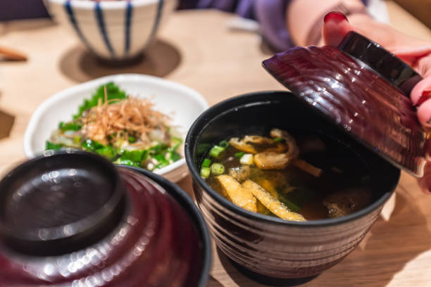 The image captures a moment of anticipation as a hand lifts the lid of a traditional Japanese ceramic bowl, revealing a steaming hot soup within. The bowl, with its dark exterior and lighter interior, cradles a clear broth garnished with green onions and likely contains ingredients such as tofu and mushrooms, suggesting a miso or dashi-based soup. The action of lifting the lid suggests the soup is being served and ready to be enjoyed. In the background, a side dish with rice and nori (seaweed), along with a blue and white striped bowl, complements the main dish, offering a balanced and traditional Japanese meal. The warm lighting and the wooden table enhance the inviting ambiance of a cozy dining experience, where the simple act of lifting the lid is a small but significant gesture in the ritual of enjoying a meal.