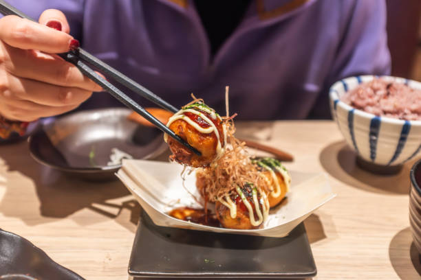 Savoring Japanese Takoyaki The image captures a culinary moment featuring a skewer of takoyaki, a popular Japanese street food. The octopus is grilled to perfection, its charred surface glistening with a glossy sheen, and it's adorned with a drizzle of mayonnaise and sprinkled with bonito flakes, adding a textural contrast and a burst of umami (savory flavor). A pair of chopsticks is poised to lift the takoyaki, ready for the diner to enjoy. In the background, a bowl of rice and a dish with a lemon wedge suggest a complete meal. The warm lighting and the focus on the food item emphasize the inviting nature of the dish, making it an irresistible treat for the senses. This scene is likely set in a restaurant that specializes in Japanese cuisine, where takoyaki is a beloved choice among patrons. cooking takoyaki is a ball shaped japanese snack stock pictures, royalty-free photos & images