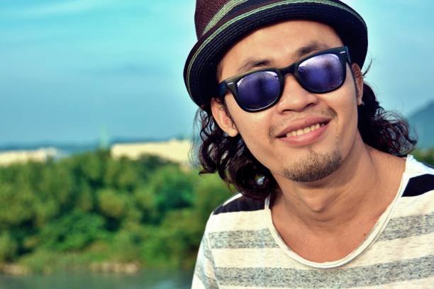 Handsome young man laughing at camera enjoying summertime day out, wearing a bucket hat and sunglasses.