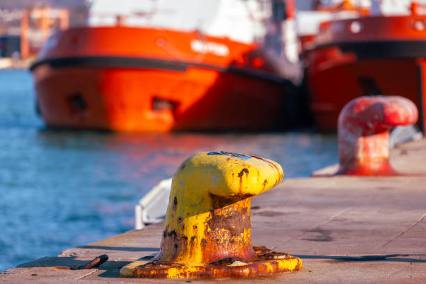 A vibrant close-up of a rusty yellow mooring bollard on a dock, with red ships anchored in the background. This image captures the essence of maritime infrastructure, harbor activity, and the shipping industry’s operational environment.