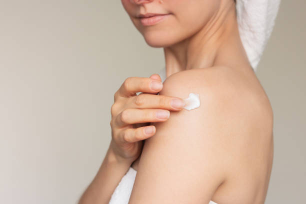 istock Woman wrapped in a white towel, touching her shoulder with her fingertips, applying a white moisturizing cream on skin. 2191050372