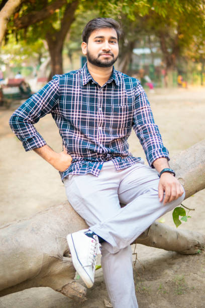 Portrait of a handsome indian young man sitting relaxed on a fallen tree and enjoying the fresh air and Tranquility of beautiful peaceful nature at day time. He is looking at the camera with a smile.