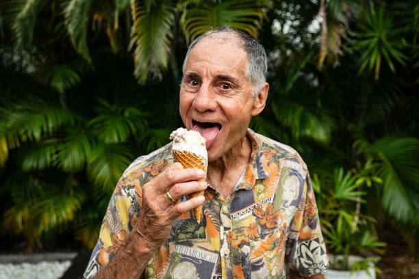 Humorous waist up view of gray-haired man in short sleeved shirt looking at camera while preparing to take a bite out of his sweet treat.
