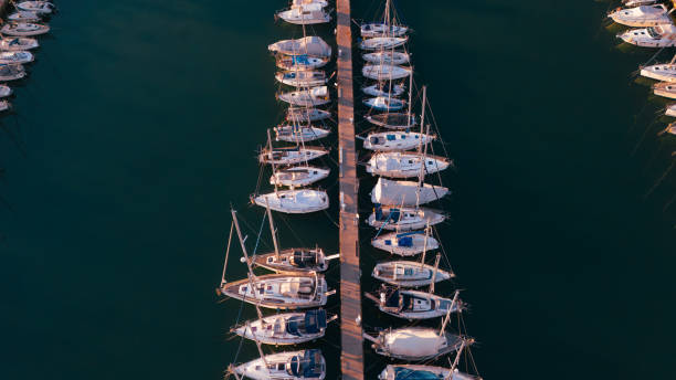 An aerial view of a marina during sunset, featuring neatly docked sailboats and yachts along wooden piers. The warm golden hues of the setting sun reflect on the calm water, evoking a peaceful and luxurious maritime atmosphere. Ideal for travel and nautical lifestyle visuals.