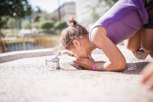 A young girl in a blue cap sits by a stone fountain, gently touching the water. Vibrant flowers cascade from the fountain, adding color to an urban setting