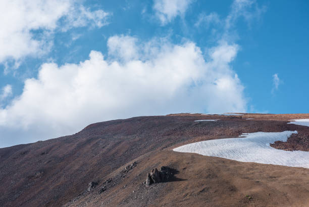 istock Rock and snowfield on mountainside in changeable weather. Sunlit stony mountain range with snow under clouds in blue sky. Sunlight and shadows on stone hill in sunny day. Rocky hillside in bright sun. 2191110483
