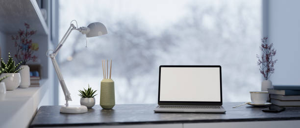 A laptop with a white desktop mockup, a table lamp, and decor on a table in a modern home office workspace, with a large window offering a view of a snowy winter landscape. 3d render, 3d illustration