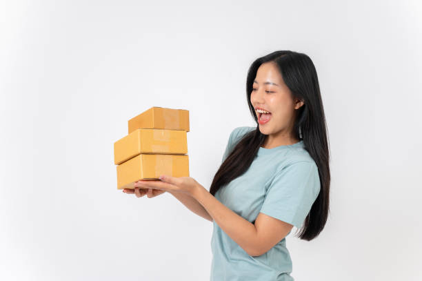 istock A cheerful Asian woman standing against a white studio background, holding cardboard boxes. 2191146414