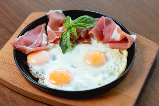 Juicy fried eggs sunny side up served with prosciutto slices and basil leaves on top in pan on wooden board, fine dining