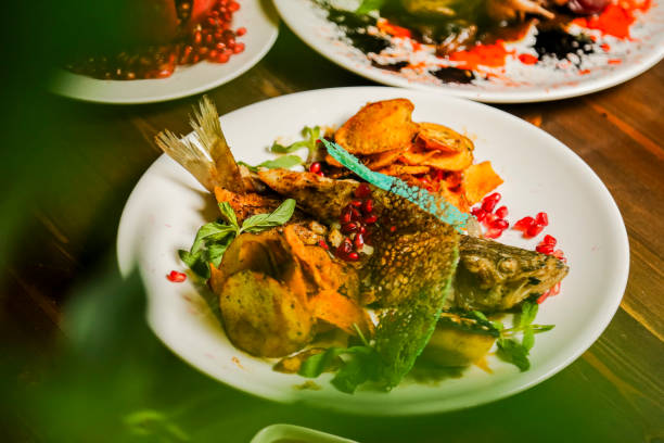 A beautifully presented dish features grilled fish topped with herbs and pomegranate seeds, accompanied by golden potato slices and colorful vegetables on a wooden table.