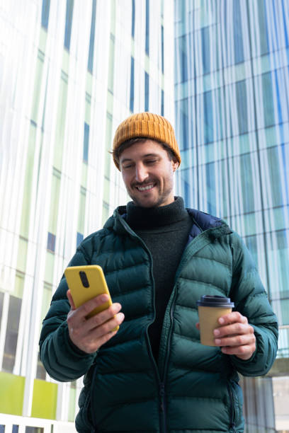 Smiling young man using smartphone and holding coffee outside office building