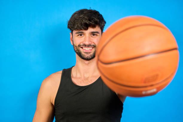 Portrait of a smiling basketball player holding a ball on a vibrant blue background, showcasing athleticism and passion for the sport