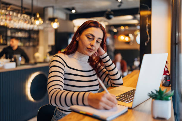 istock Focused female entrepreneur works from coffee shop, arranges paperwork, transcribes important information from laptop screen into her notebook 2191171797