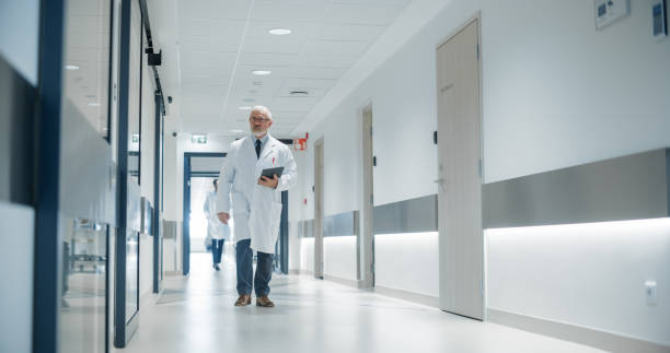istock Portrait of a Senior General Practitioner Using Tablet Computer to as He is Walking and Browsing Patient's Medical History. Hospital Personnel Walking Through a Modern Clinic Corridor 2191183074