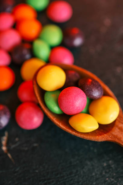 Colorful candy-coated chocolates in a wooden spoon on a dark textured surface. Bright, vibrant, and sweet treats with a fun and playful arrangement, perfect for dessert or snack concepts.