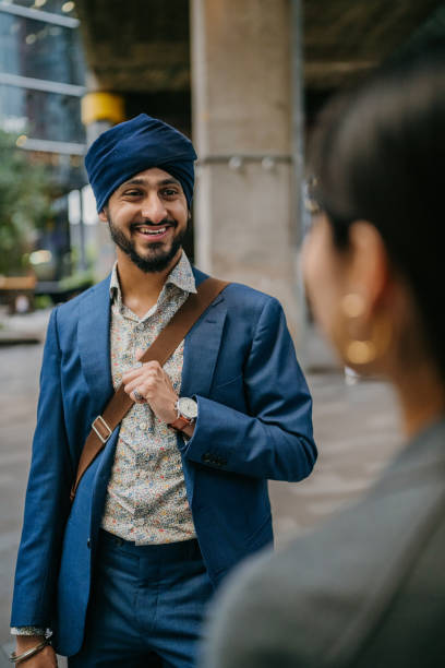 A stylish businessman with a blue turban and floral shirt smiles warmly, standing in a vibrant city setting, showcasing contemporary style and optimism.