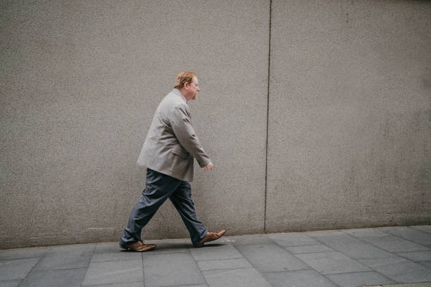 istock Figure in grey walks a city path, wall beside him. 2191194020
