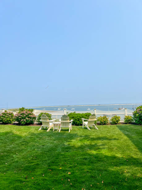 istock A serene backyard with white Adirondack chairs, lush green grass, blooming hydrangeas, a white picket fence, and a view of calm waters dotted with boats under a clear blue sky. 2191196564