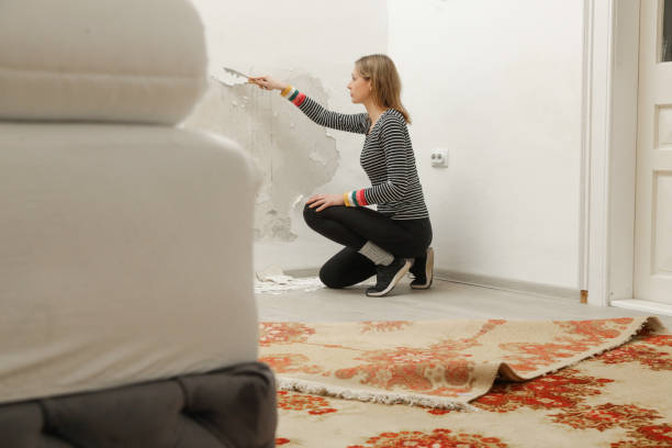istock Saltpeter or mold on the wall problem. Woman is using a scraper to scrape and remove all loose paint and plaster that is in poor condition, until a firm surface is achieved. 2191197395