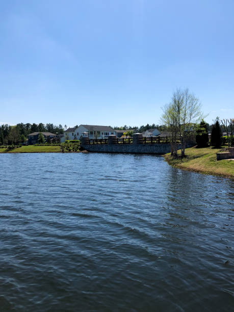 istock A calm lake stretches under a clear blue sky, bordered by grassy banks and a stone-lined bridge, with houses and trees in the distance, creating a peaceful suburban waterside scene. 2191199265