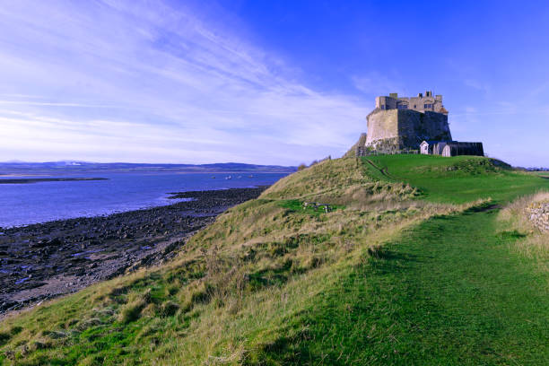 A view of Lindisfarne Castle on the Holy Isle in Northumberland, England against a pale summer sky with headland and boats in the foreground.