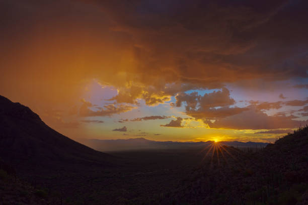 A breathtaking sunset scene featuring a desert valley, silhouetted mountains, and brilliant orange and yellow hues in the dramatic sky, creating a serene and awe-inspiring nature landscape.