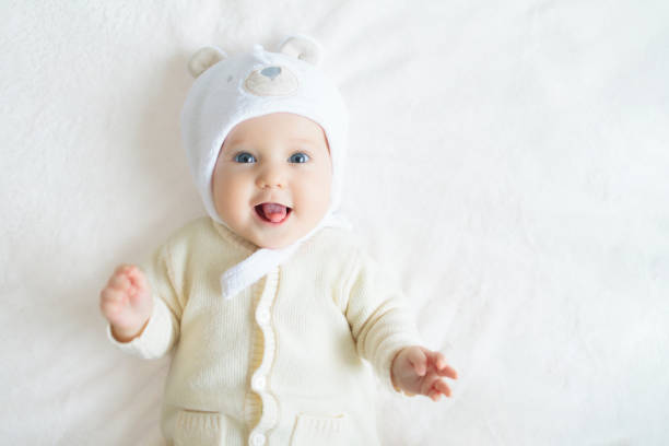 Adorable baby in hat in white sunny bedroom. Newborn child relaxing in bed. Nursery for young children. Winter clothing for kids. Family morning at home. New born kid during tummy time with toy bear.