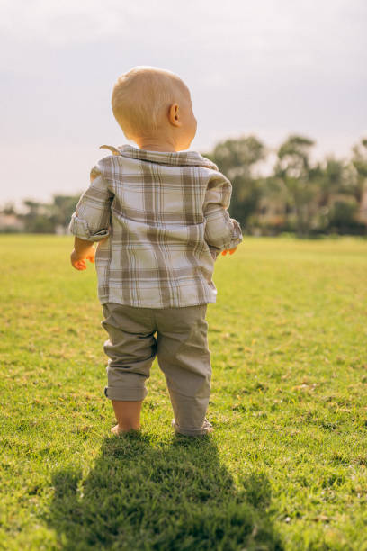 A tender photograph capturing the back view of a baby standing confidently on a sunlit grassy field. The soft plaid shirt and rolled-up pants add charm to the moment, as the baby gazes toward the horizon, symbolizing curiosity and the start of endless adventures. The serene park and gentle sunlight create a backdrop of tranquility and growth.