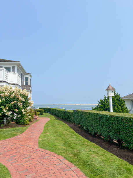 istock A charming coastal walkway with a red brick path winds past manicured hedges, blooming flowers, and a birdhouse, leading to a serene seaside view under a bright blue sky. 2191222068