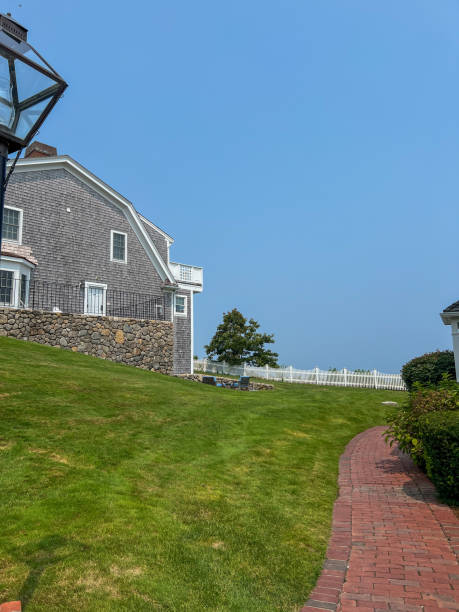 istock A classic coastal home with stone and shingle siding sits beside a red brick path and a lush green lawn, with a white picket fence and a clear blue sky completing the serene outdoor scene. 2191222073
