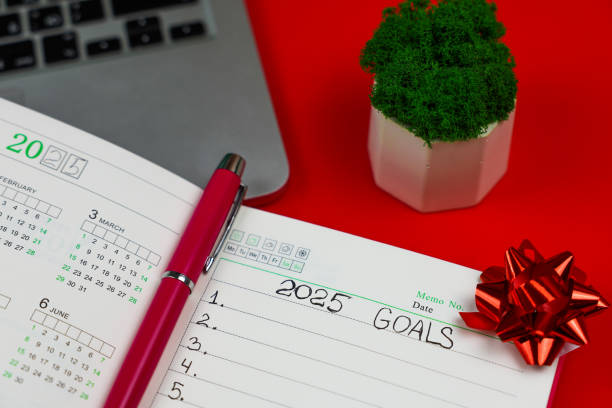 istock Close-up of a pink notebook diary in which it is written: Goals for 2025, inside it is a calendar for 2025 and there is a pink pen and in the upper right corner there is a festive New Year's bow on a red background, an open laptop lies on top, part of the 2191224876