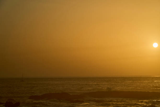 small sailing boat cruising across the deep blue atlantic ocean along the rugged and rocky coastline of Portugal during a beautiful golden sunset.