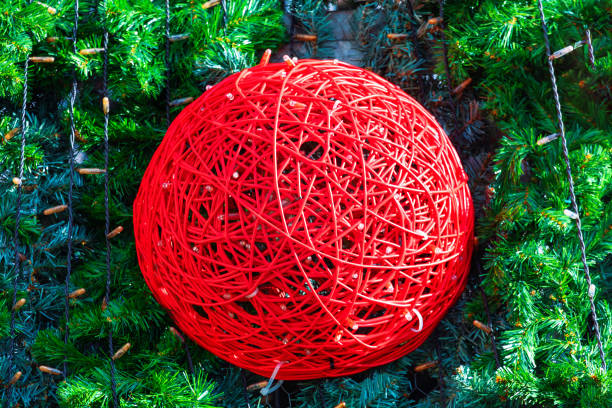 Spherical decoration made of red intertwined wires, set against a backdrop of green pine branches. Red sphere stands out vividly due to its bright color and intricate design. Decoration is part of a Christmas celebration