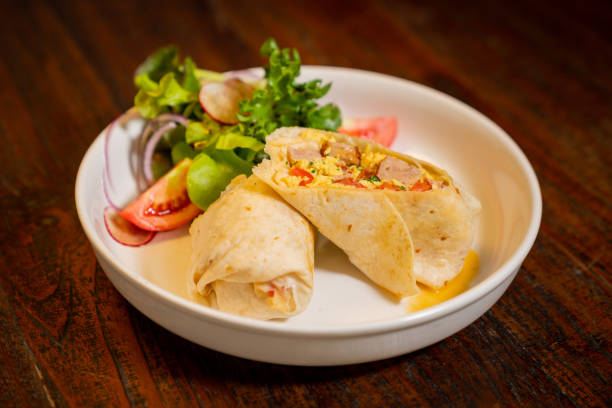 istock Rolled tortillas filled with various ingredients on plate, includes green leaves, tomato slices, and vegetables, the backdrop is a wooden table 2191245869