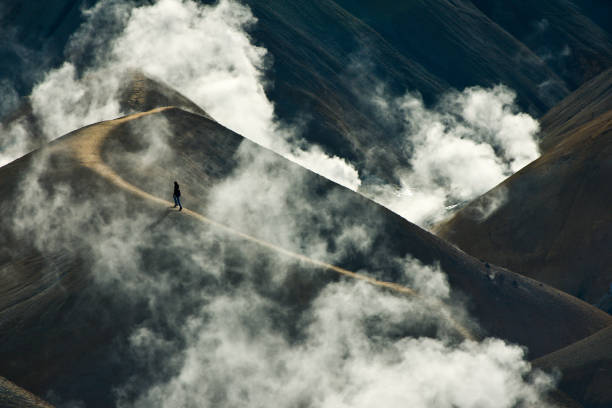 Lone female hiker ascends a steep slope through rising steam clouds in a geothermal landscape in the Kjolur Interior Route central Iceland, only open mid-late summer.