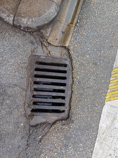 istock Storm drain embedded in a weathered asphalt road, surrounded by damp patches and worn curbstones with a hint of yellow marking. 2191279710