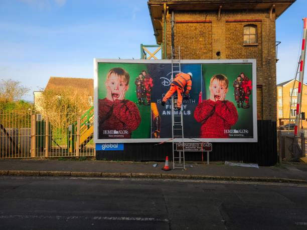 Crawley, UK - 20 December, 2024: a worker wearing high visibility clothing is up a ladder removing a billboard poster in Crawley, UK. The poster is advertising the Home Alone films streaming on the Disney Plus platform.