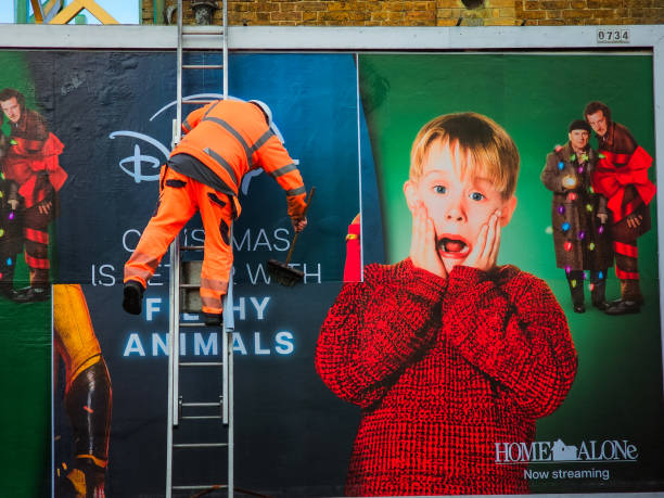 Crawley, UK - 20 December, 2024: a worker wearing high visibility clothing is up a ladder removing a billboard poster in Crawley, UK. The poster is advertising the Home Alone films streaming on the Disney Plus platform.