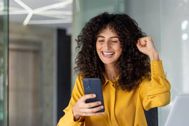 istock Excited woman celebrating success while using smartphone in modern office space, expressing happiness and achievement 2191291592